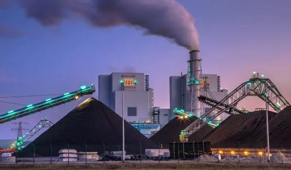 An industrial plant with a smoking chimney and conveyor belts is seen during twilight, with piles of black material in the foreground.