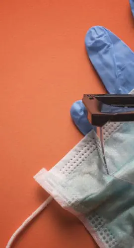 A pair of eyeglasses sits on top of a face mask and blue medical gloves against an orange background.