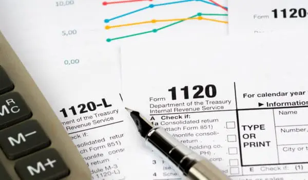 A calculator and a pen are placed on top of scattered IRS tax forms, including Form 1120 and 1120-L, with graphs in the background.
