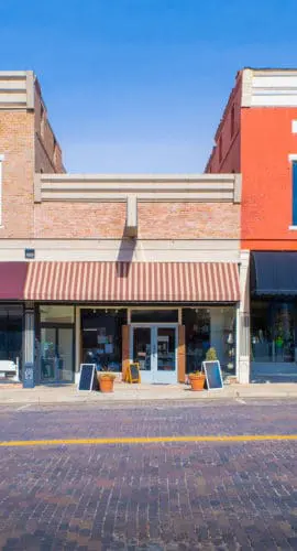 Two storefronts, one with a red brick facade and the other with a light brown brick facade, are visible along a paved street.