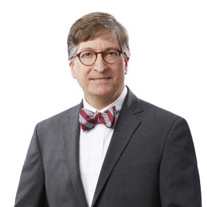 A man wearing a bowtie and glasses is smiling against a plain background.